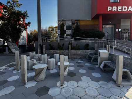 Presbeton H-E-X,Vonkajšie posedenie s geometrickými kamennými stolmi a stoličkami je krásne postavené na pozadí zámkovej dlažby, obklopené nízkou stenou a stromami, výhodne umiestnené v blízkosti vchodu do budovy.
